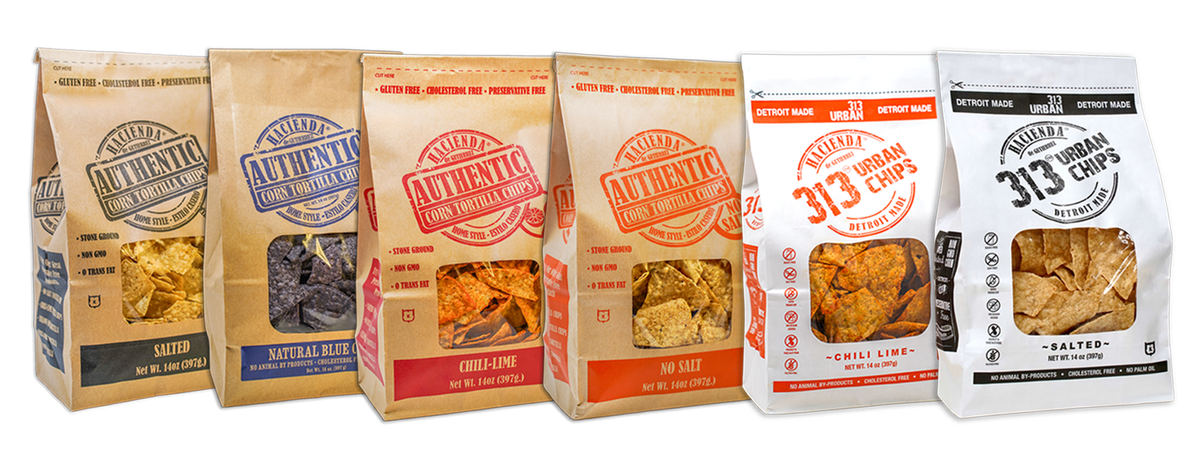 A display featuring six bags of chips. The first four bags are Hacienda Authentic Corn Tortilla Chips. The first bag is labeled "Salted," the second bag is "Natural Blue," the third bag is labeled "Chili-Lime," and the fourth bag is labeled "No Salt." Each of these bags is brown and features a transparent window to view the contents. The last two bags are Hacienda 313 Urban Chips, with the first labeled "Chili Lime" in red, followed by a "Salted" in black. All bags prominently display "DETROIT MADE" and fea