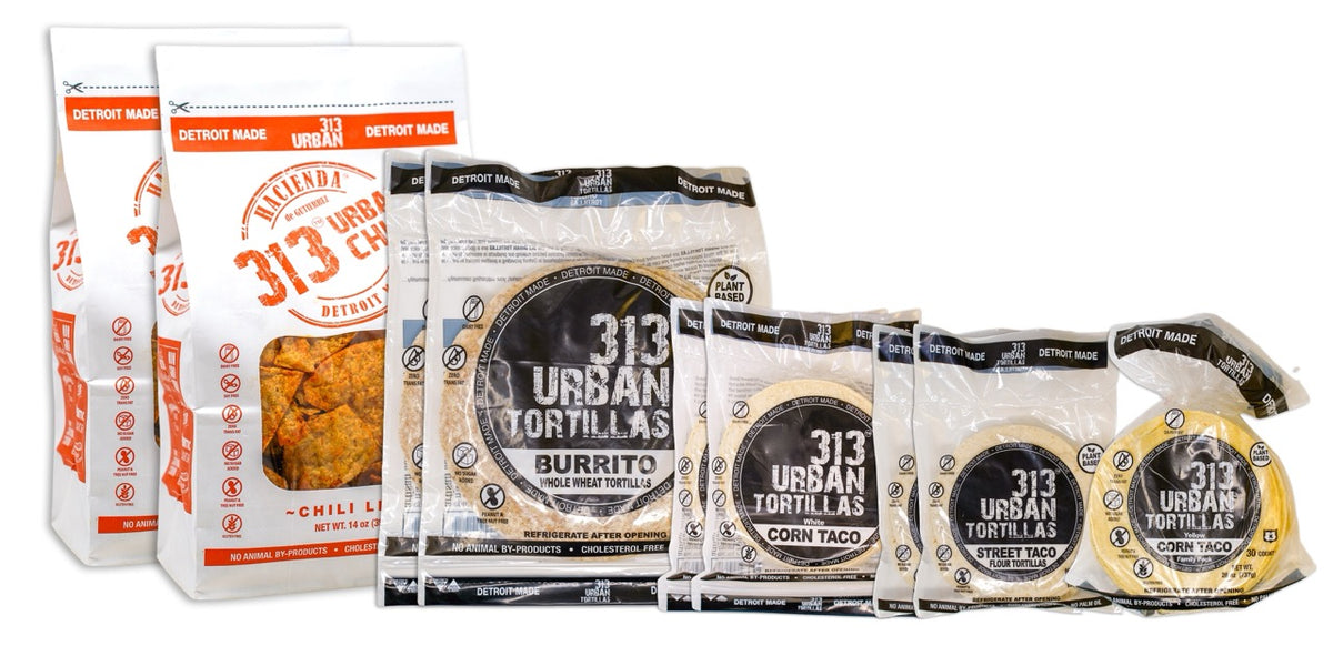 A display featuring several Hacienda 313 Urban products. At the forefront are two bags of Chili Lime tortilla chips, labeled &quot;313 Urban Chips&quot; with vibrant red labeling. Behind them are two bags of 8&quot; Whole Wheat tortillas, labeled &quot;313 Urban Tortillas,&quot; followed by two bags of White Corn tortillas, also labeled &quot;313 Urban Tortillas.&quot; Next, there are two bags of 5&quot; Flour tortillas labeled &quot;313 Urban Tortillas,&quot; and finally, a single bag of Yellow Corn tortillas, completing the assortment. All bags prominent