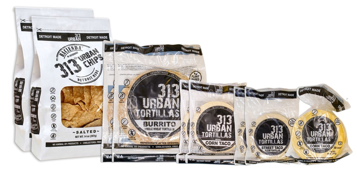 A display featuring several Hacienda 313 Urban products. At the forefront are two bags of Salted tortilla chips, labeled &quot;313 Urban Chips&quot; with black labeling. Behind them are two bags of 8&quot; Whole Wheat tortillas, labeled &quot;313 Urban Tortillas,&quot; followed by two bags of White Corn tortillas, also labeled &quot;313 Urban Tortillas.&quot; Next, there are two bags of 5&quot; Flour tortillas labeled &quot;313 Urban Tortillas,&quot; and finally, a single bag of Yellow Corn tortillas, completing the assortment. All bags prominent