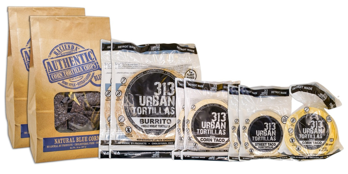 A display featuring several Hacienda products. At the forefront are two bags of Authentic Natural Blue Corn tortilla chips, labeled &quot;Hacienda Authentic Corn Tortilla Chips&quot; in brown packaging. Behind them are two bags of 8&quot; Whole Wheat 313 Urban Tortillas, followed by two bags of White Corn 313 Urban Tortillas. Next, there are two bags of 5&quot; Flour 313 Urban Tortillas, and finally, a single bag of Yellow Corn 313 Urban Tortillas. All bags prominently feature &quot;DETROIT MADE&quot; on the packaging.