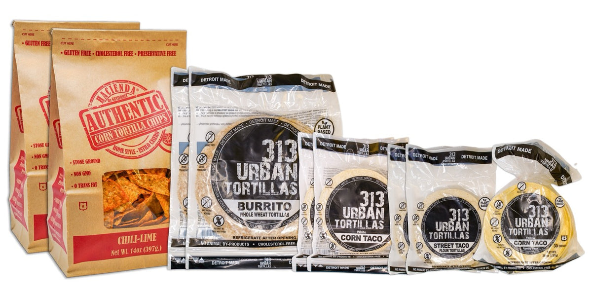 A display featuring several Hacienda products. At the forefront are two bags of Authentic Chili Lime tortilla chips, labeled &quot;Hacienda Authentic Corn Tortilla Chips&quot; in brown packaging. Behind them are two bags of 8&quot; Whole Wheat 313 Urban Tortillas, followed by two bags of White Corn 313 Urban Tortillas. Next, there are two bags of 5&quot; Flour 313 Urban Tortillas, and finally, a single bag of Yellow Corn 313 Urban Tortillas. All bags prominently feature &quot;DETROIT MADE&quot; on the packaging.