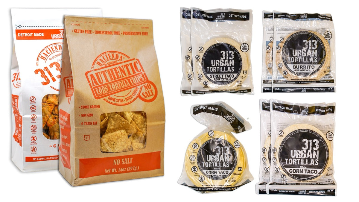 A display featuring several Hacienda products. At the forefront are one bag of Hacienda Authentic No Salt tortilla chips and one bag of 313 Chili Lime tortilla chips, both labeled prominently. Behind them are two bags of 8&quot; Whole Wheat 313 Urban Tortillas. Next are two bags of White Corn 313 Urban Tortillas, followed by two bags of 5&quot; Flour 313 Urban Tortillas. Finally, there is one bag of Yellow Corn 313 Urban Tortillas. All bags prominently feature &quot;DETROIT MADE&quot; on their packaging.
