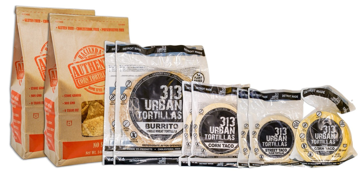 A display featuring several Hacienda products. At the forefront are two bags of Authentic No Salt tortilla chips, labeled &quot;Hacienda Authentic Corn Tortilla Chips&quot; in brown packaging. Behind them are two bags of 8&quot; Whole Wheat 313 Urban Tortillas, followed by two bags of White Corn 313 Urban Tortillas. Next, there are two bags of 5&quot; Flour 313 Urban Tortillas, and finally, a single bag of Yellow Corn 313 Urban Tortillas. All bags prominently feature &quot;DETROIT MADE&quot; on the packaging.